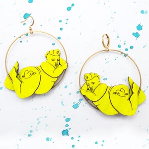 May include: A pair of gold hoop earrings featuring a yellow cartoon figure of a person lying down. The figure has a heart with an arrow on its backside.