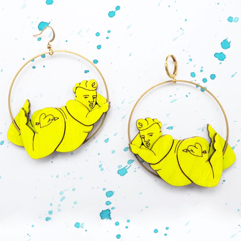 May include: A pair of gold hoop earrings featuring a yellow cartoon figure of a person lying down. The figure has a heart with an arrow on its backside.