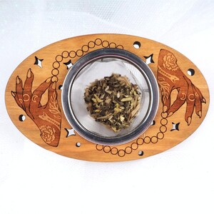 May include: Wooden tea infuser with a stainless steel mesh basket, holding loose leaf tea. The wooden base features etched hands, crescent moons, stars, and a beaded chain design. The infuser is oval-shaped.