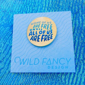 May include: A wooden pin with the text "NONE OF US ARE FREE UNTIL ALL OF US ARE FREE" in blue and gold. The pin is on a light blue card with the words "WILD FANCY DESIGN" printed on it.