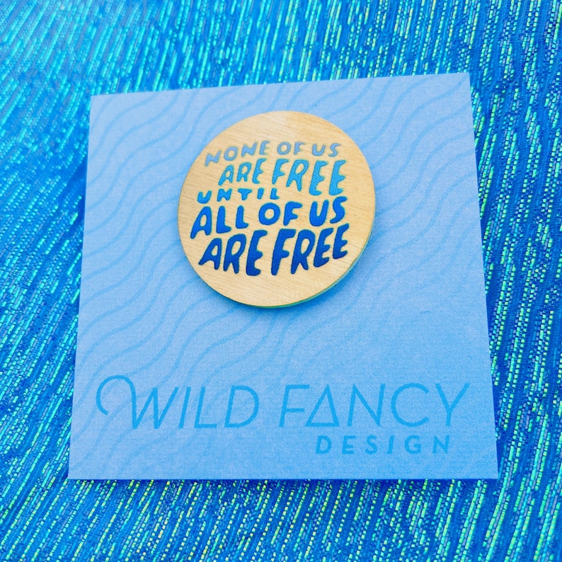 May include: A wooden pin with the text "NONE OF US ARE FREE UNTIL ALL OF US ARE FREE" in blue and gold. The pin is on a light blue card with the words "WILD FANCY DESIGN" printed on it.