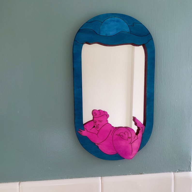 Puede incluir: Un espejo de madera azul con un dise&ntilde;o tallado de una ola y una luna. El espejo refleja una figura rosa de una persona con un dise&ntilde;o de ola.