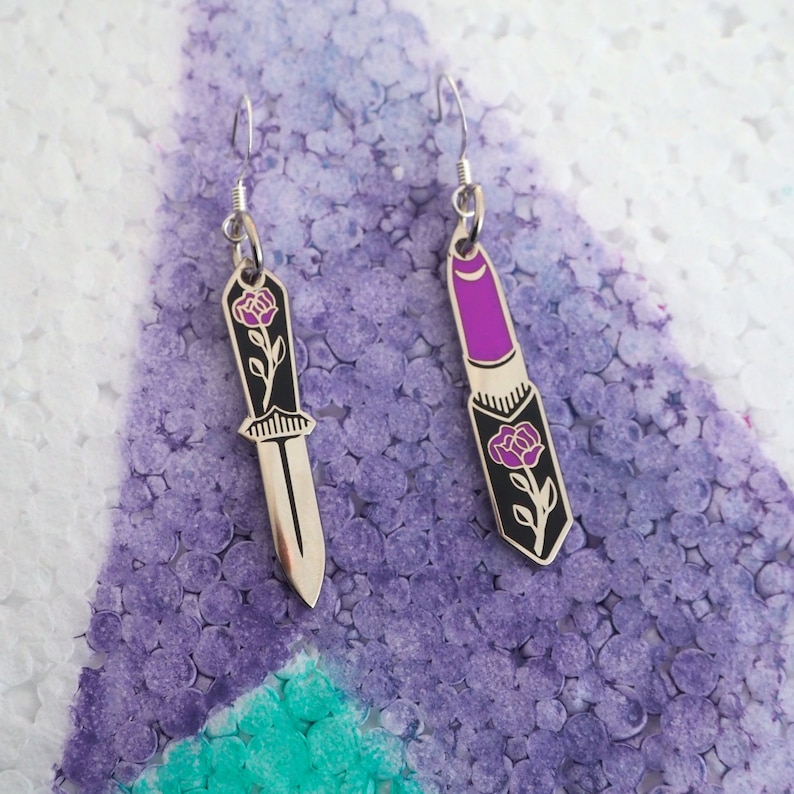 May include: A pair of earrings featuring a silver knife with a purple rose and a silver lipstick with a purple rose.
