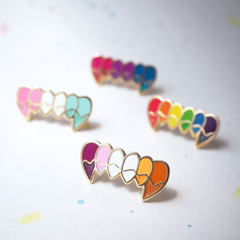 May include: Set of five enamel pins shaped like hearts, each with a different color: purple, pink, white, orange, and red. The pins are arranged in a row, with the purple heart on the left and the red heart on the right.