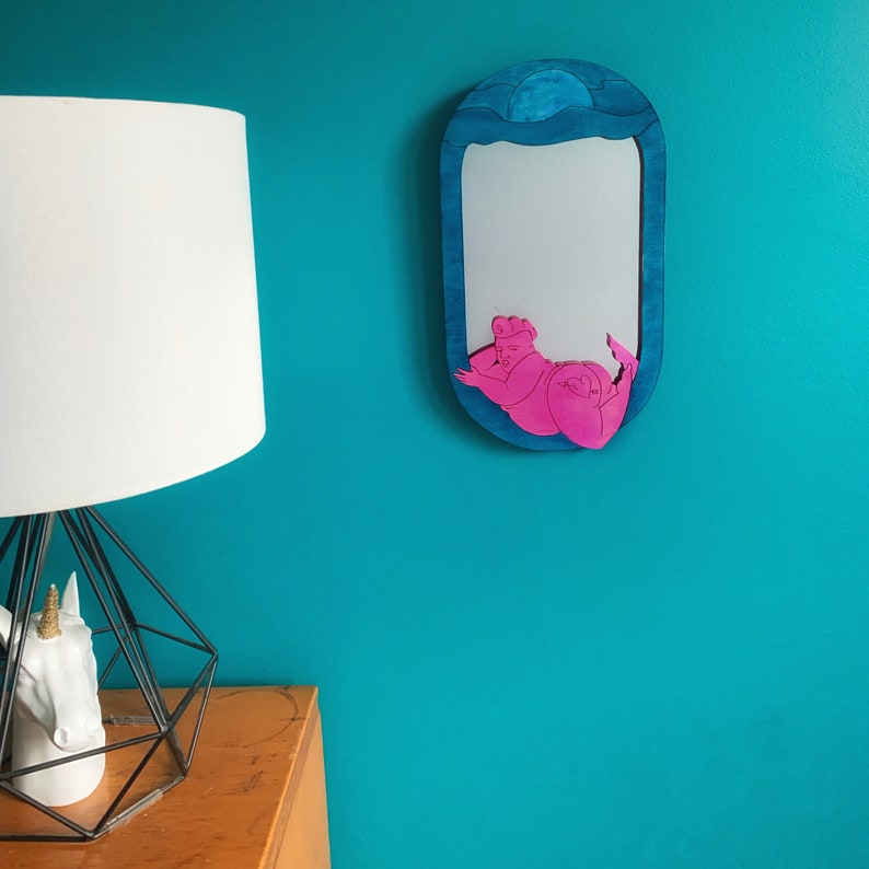 Puede incluir: Un espejo ovalado azul con una silueta rosa de una persona tumbada boca abajo con los brazos extendidos. El espejo est&aacute; colgado en una pared turquesa.