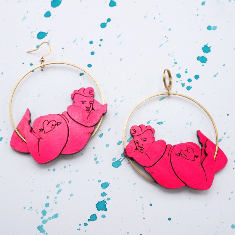 May include: A pair of gold hoop earrings featuring pink wooden figures of people. The figures are in a relaxed pose with their arms outstretched.
