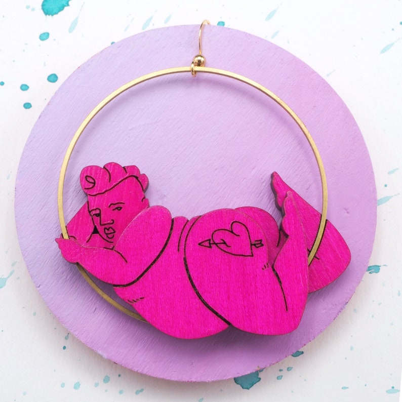 May include: Pink wooden earring with a gold hoop and a cutout of a person with a heart tattoo on their hip.