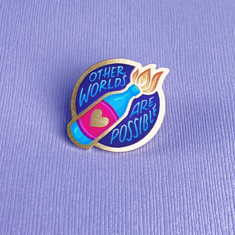 May include: A round pin with a blue bottle design, a pink heart, and the words "OTHER WORLDS ARE POSSIBLE" in a gold frame. The bottle has a flame coming out of the top. The pin is on a purple background.