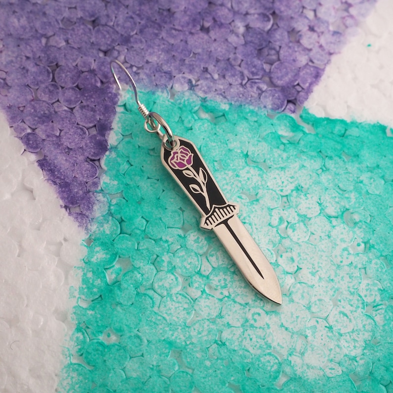 May include: A silver earring shaped like a knife with a black handle and a pink rose design.