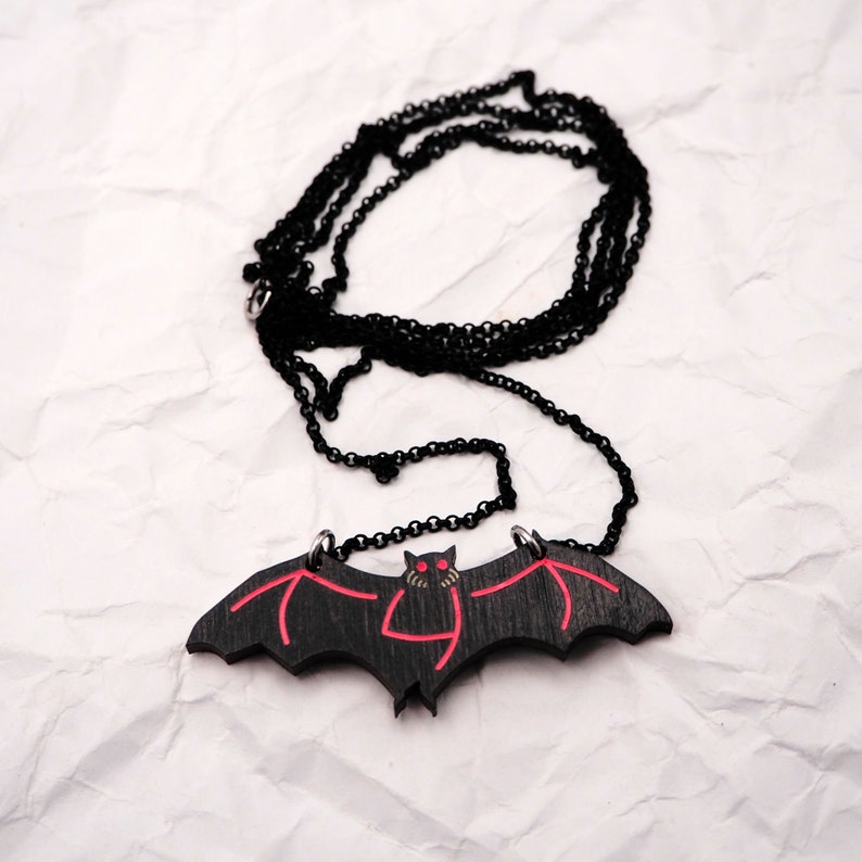 May include: A black bat pendant necklace with red eyes and wings. The necklace is on a black chain.