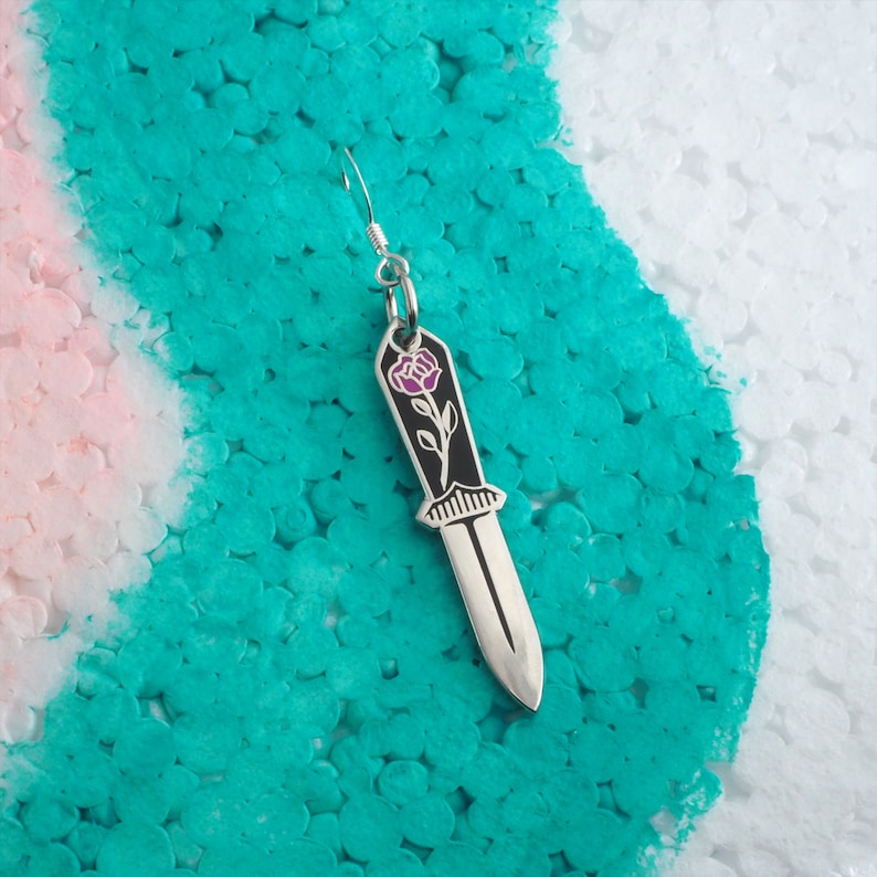 May include: Silver earring with a black dagger charm featuring a pink rose design.