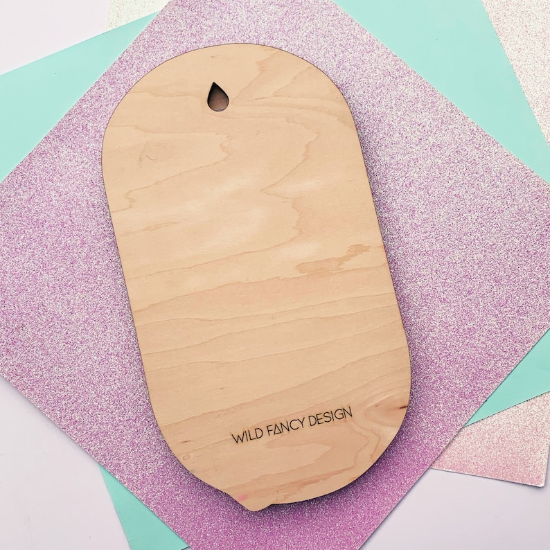 Puede incluir: Un letrero ovalado de madera clara con un agujero en la parte superior para colgar. El letrero tiene el texto "WILD FANCY DESIGN" impreso en la parte inferior.