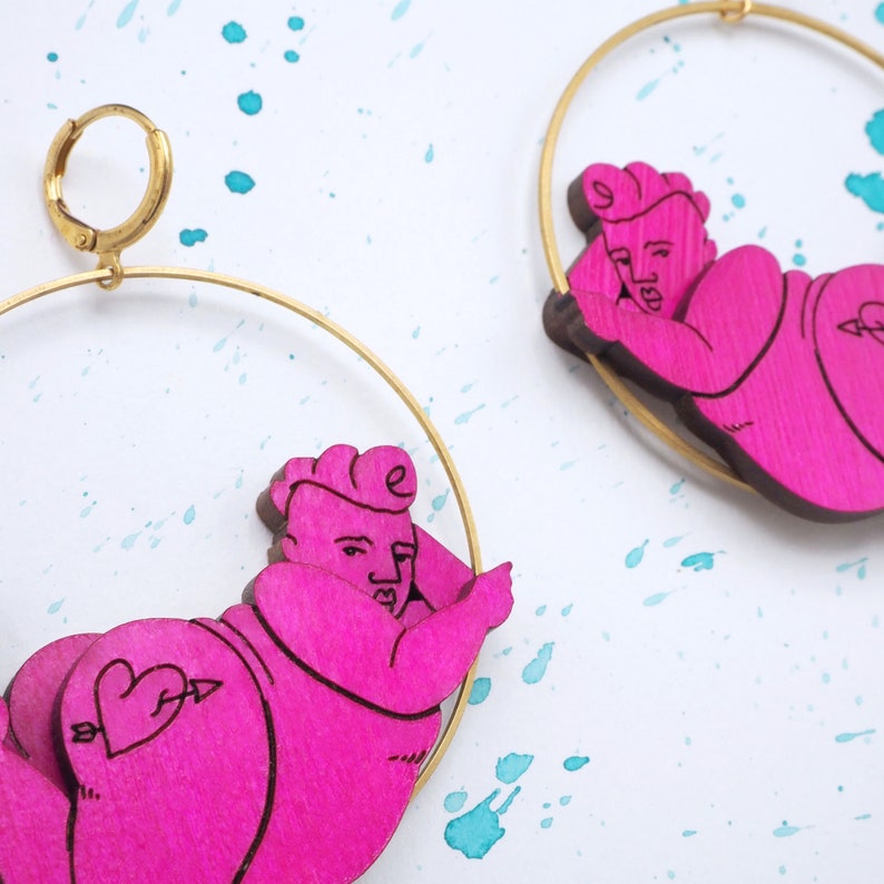 May include: A pair of pink wooden earrings with a gold hoop. Each earring features a cartoon figure of a person with a heart on their backside.