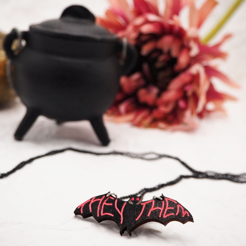 May include: A black wooden bat necklace with red lettering that says "they/them". The necklace is on a white surface with a black cauldron and a pink flower in the background.
