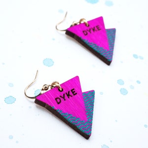 May include: A pair of pink triangle earrings with the word "DYKE" engraved in black lettering. The earrings have a teal and pink geometric pattern.