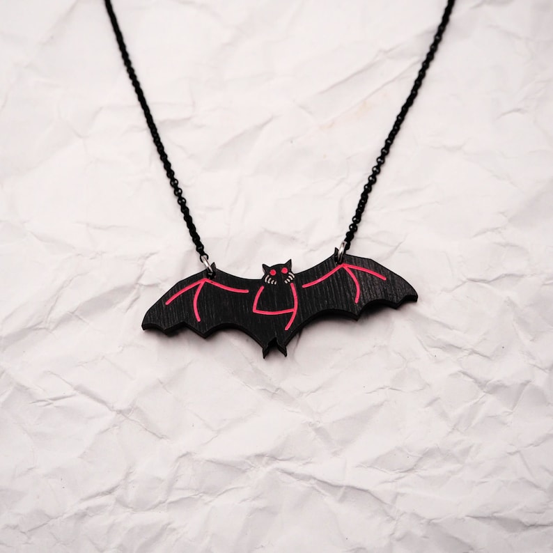 May include: A black bat pendant necklace with red detailing. The bat has red eyes and a red mouth. The necklace is on a white crumpled background.