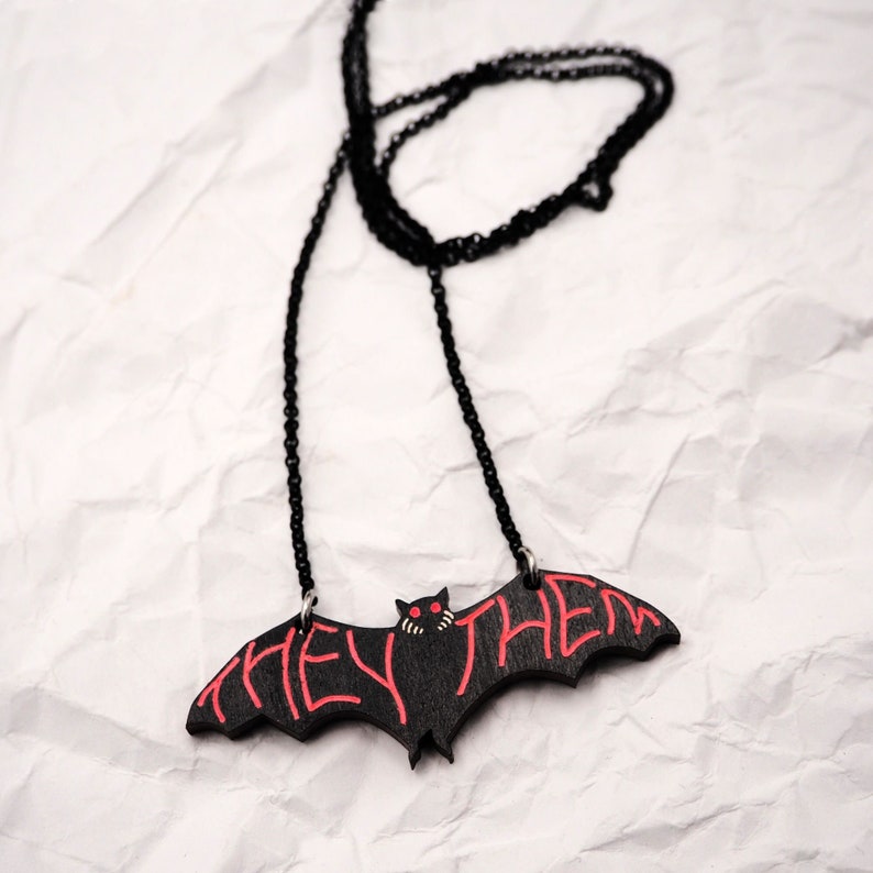 May include: A black bat pendant necklace with a red "they/them" inscription. The necklace has a black chain.