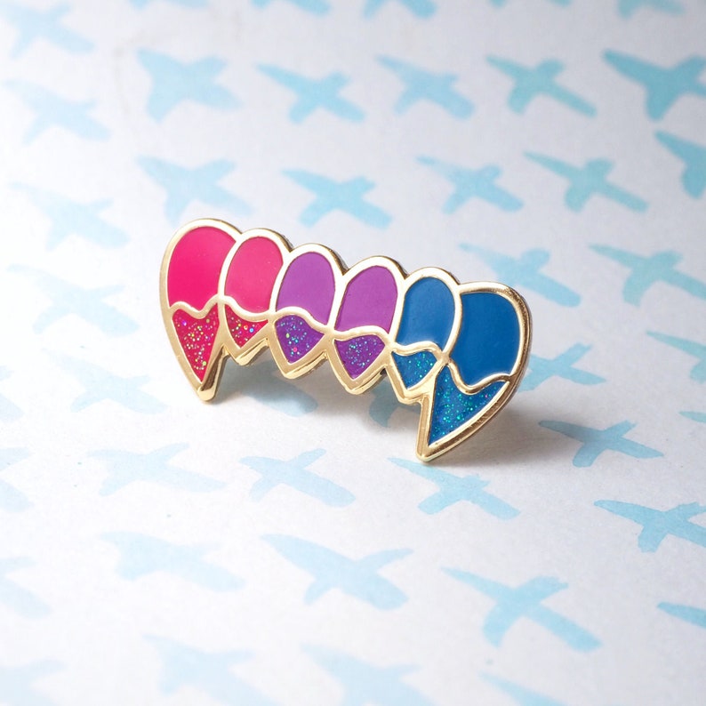 May include: A gold-toned enamel pin with five hearts in a row. The hearts are pink, red, purple, light purple, and blue. Each heart has glitter inside.