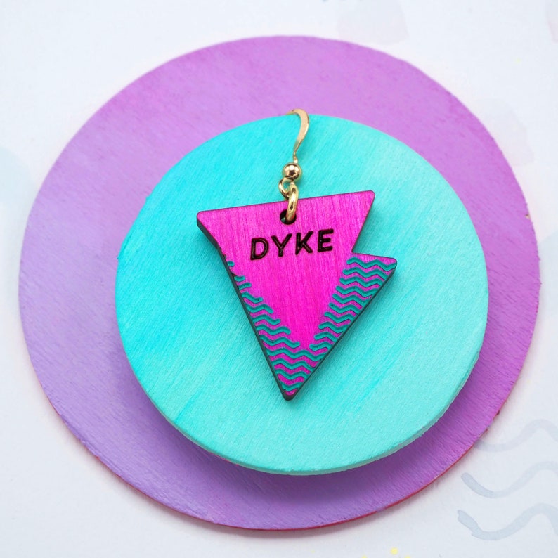 May include: Pink triangle-shaped wooden earring with the word "DYKE" written in black lettering. The earring is hanging from a gold hook and is on a background of light purple and turquoise.