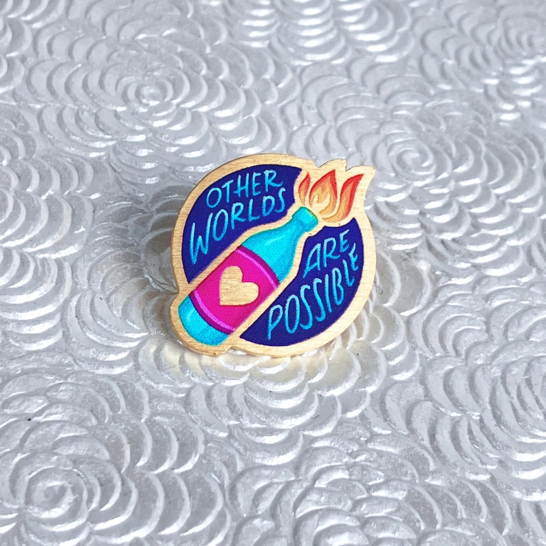 May include: A round wooden pin with a blue border and the words "OTHER WORLDS ARE POSSIBLE". The pin features a blue bottle with a pink heart and a flame. The pin is set against a white, textured background.