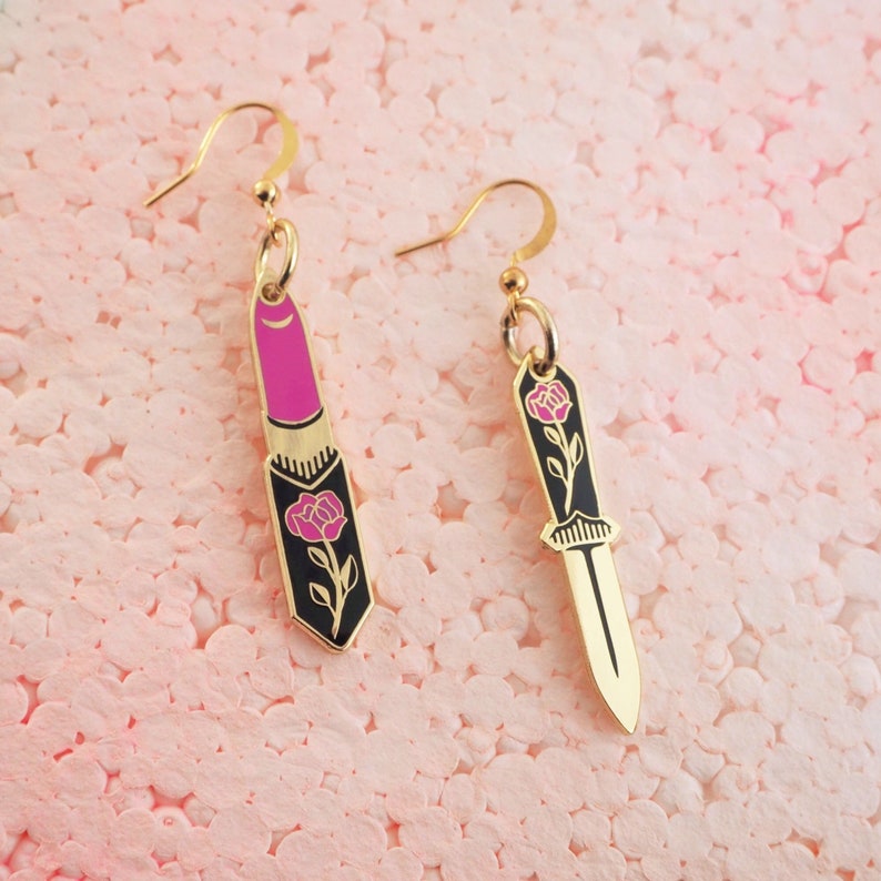 Op de afbeelding: Een paar gouden oorbellen met een ontwerp van roze lipstick en een zwart mes. Elke oorbel is versierd met een roze roos en een groene steel.