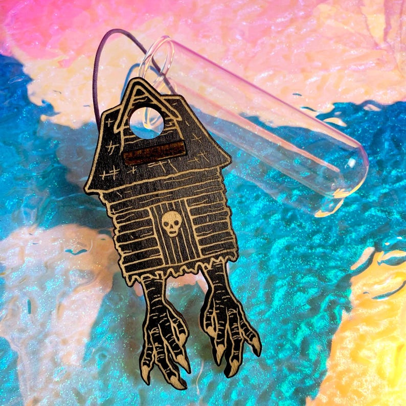 May include: A wooden house-shaped charm with a skull on the door and bird feet. The charm is black with a light brown wood grain. A clear glass tube is attached to the charm. The background is a mix of blue, pink, and yellow.