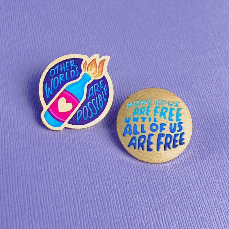 May include: Two enamel pins on a purple surface. One pin features a blue bottle with a heart and flame, surrounded by text that reads "OTHER WORLDS ARE POSSIBLE." The other pin is gold with blue text: "NONE OF US ARE FREE UNTIL ALL OF US ARE FREE."
