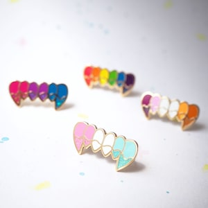 May include: Set of five enamel pins shaped like fangs, each with a different color gradient. The colors include pink, white, blue, red, orange, yellow, and purple.