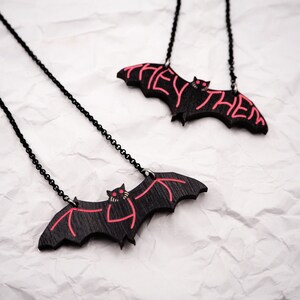 May include: Two black bat necklaces with red painted details. The bats have the words "They Them" written on them.