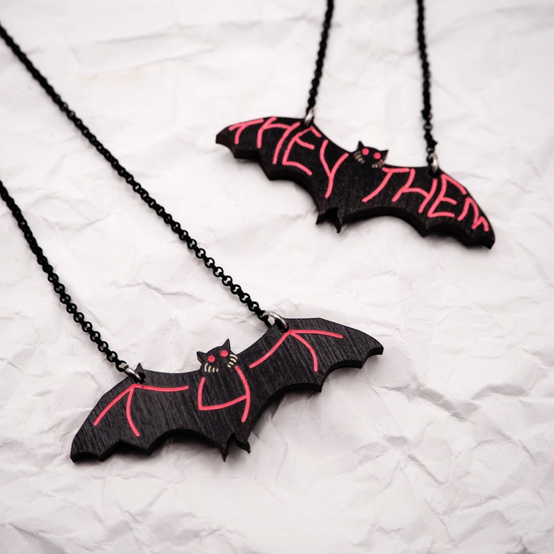 May include: Two black bat necklaces with red painted details. The bats have the words "They Them" written on them.