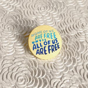 May include: A round, gold-colored pin with the text "NONE OF US ARE FREE UNTIL ALL OF US ARE FREE" in blue and light blue lettering. The pin is set against a textured, white background.