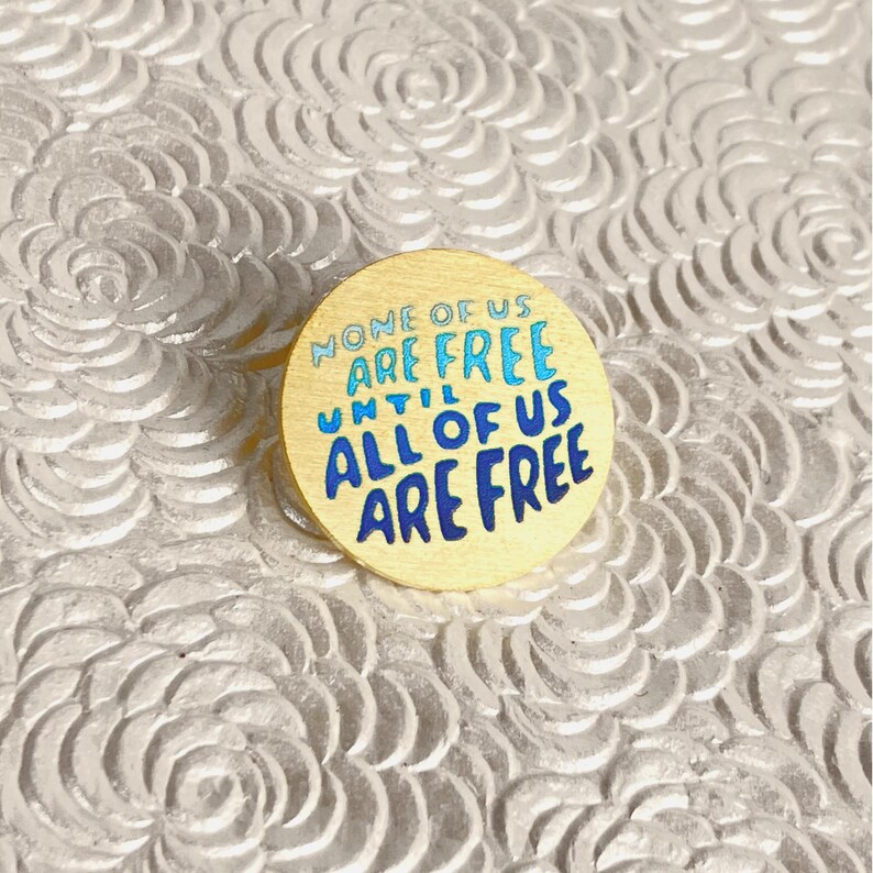 May include: A round, gold-colored pin with the text "NONE OF US ARE FREE UNTIL ALL OF US ARE FREE" in blue and light blue lettering. The pin is set against a textured, white background.