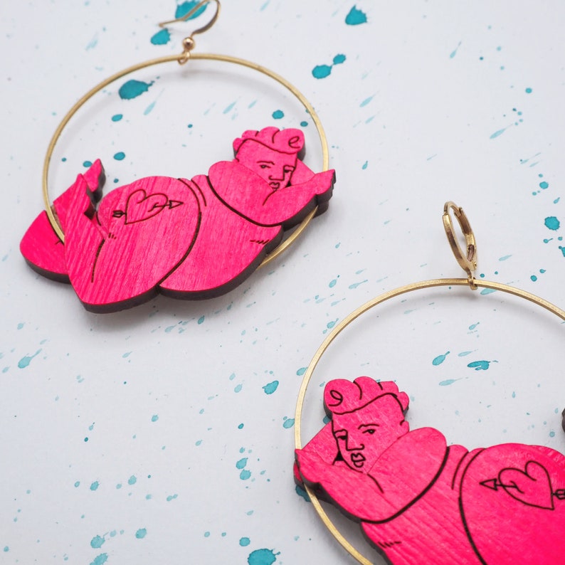 May include: A pair of gold hoop earrings with pink wooden cutout figures of a person lying down. The figures have a heart with an arrow through it on their back.