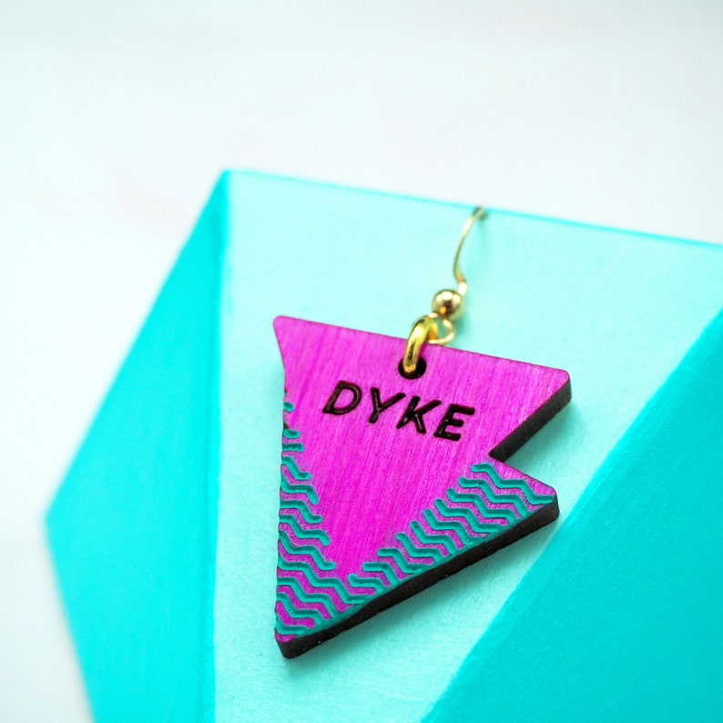 May include: Pink triangle earring with a gold hook and the word "DYKE" written in black lettering. The triangle has a turquoise and black geometric pattern.