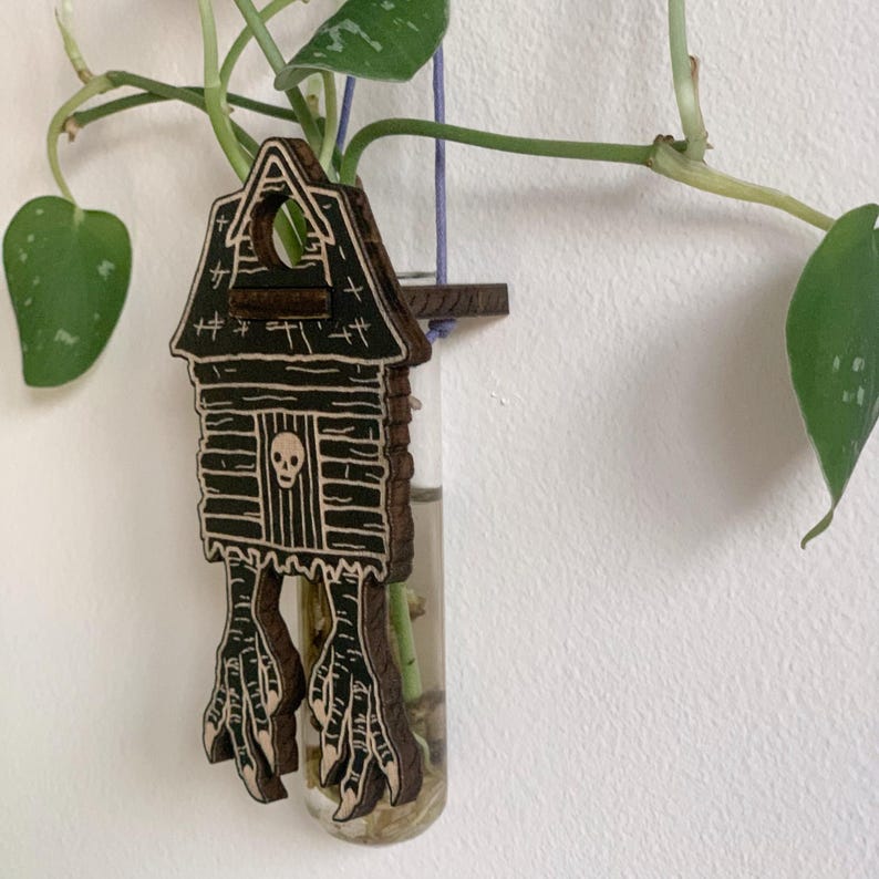 May include: A wooden house-shaped propagation station with a skull on the door and claw feet. The house holds a clear glass tube for a plant cutting. The house is brown with black etched details and is hanging on a wall with a green plant.