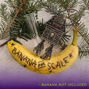 May include: A wooden ornament shaped like a house with bird feet, a skull, and a purple loop for hanging. A banana with the words "Banana for Scale" is in the foreground. A clear glass and pine branches are in the background. The text "Banana Not Included" is at the bottom.