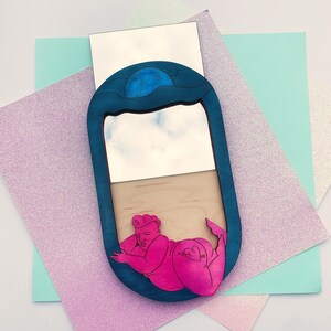 Puede incluir: Un espejo ovalado azul y blanco con una figura rosa de una persona tumbada en su interior. El espejo tiene un dise&ntilde;o de ola azul en la parte superior y un marco de madera.