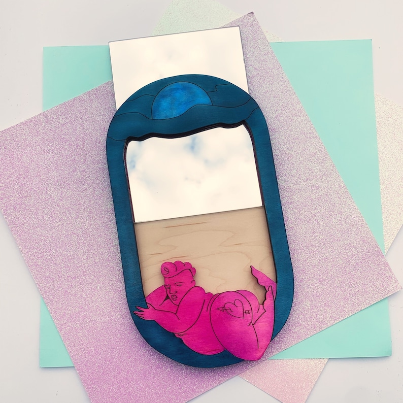 Puede incluir: Un espejo ovalado azul y blanco con una figura rosa de una persona tumbada en su interior. El espejo tiene un dise&ntilde;o de ola azul en la parte superior y un marco de madera.