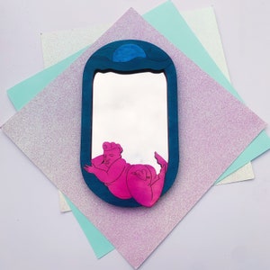 Puede incluir: Un espejo de madera azul y rosa con una ilustraci&oacute;n estilizada de una persona tumbada. El espejo tiene una forma redondeada y un marco azul.