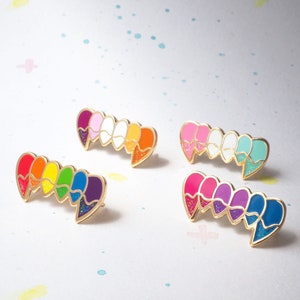 May include: Set of four enamel pins featuring a rainbow gradient of colors in the shape of a heart. Each pin has a gold metal backing.