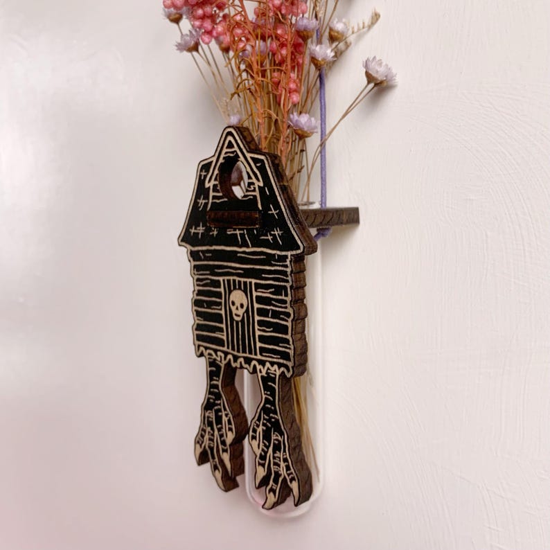May include: A wooden, Halloween-themed vase with a house design, featuring a skull and bird feet. The vase holds dried pink and white flowers. The vase is attached to a clear glass tube.
