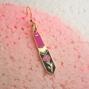 Op de afbeelding: Gouden oorbel met een roze lipstick-bedel met een zwart rozenontwerp.