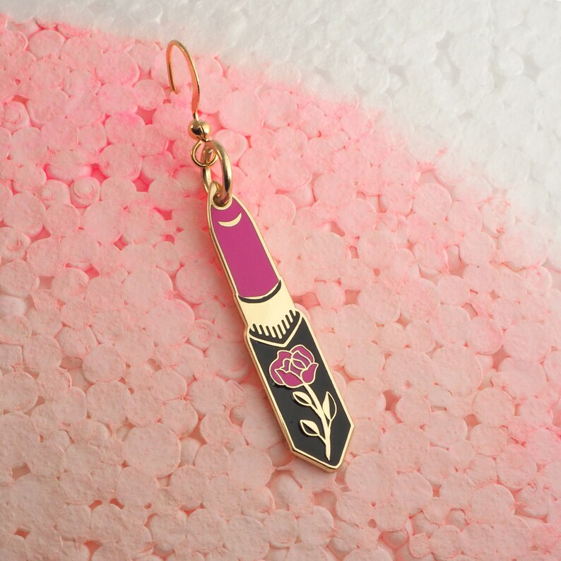 Op de afbeelding: Gouden oorbel met een roze lipstick-bedel met een zwart rozenontwerp.