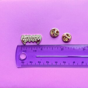 May include: A silver enamel pin with the text "WILD FANCY DEPT" in a gothic font. The pin is shaped like a set of fangs and is about 1 inch long. The pin is on a purple surface with a ruler for scale.