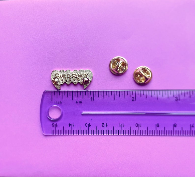 May include: A silver enamel pin with the text "WILD FANCY DEPT" in a gothic font. The pin is shaped like a set of fangs and is about 1 inch long. The pin is on a purple surface with a ruler for scale.