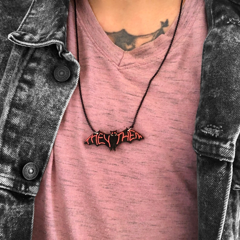 May include: A black bat-shaped necklace with the words "they/them" in pink lettering.