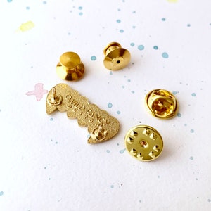 May include: Gold-colored enamel pin with the words "Wild & Free" in a heart shape. The pin has a butterfly design and comes with a safety clasp.