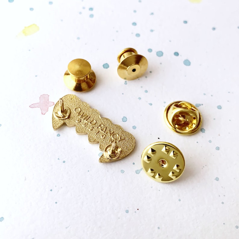 May include: Gold-colored enamel pin with the words "Wild & Free" in a heart shape. The pin has a butterfly design and comes with a safety clasp.