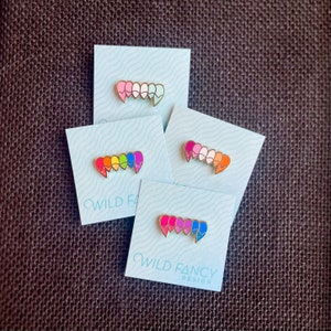May include: Four enamel pins shaped like a set of fangs, each with a different color gradient. The pins are on white cards with the text "WILD FANCY DESIGN" on the bottom.
