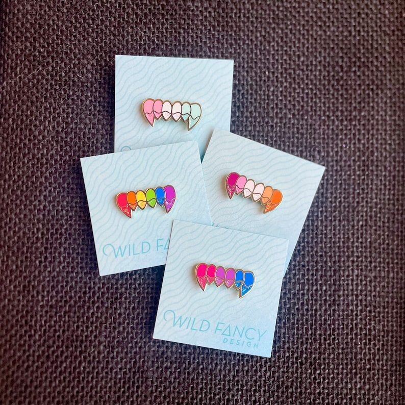 May include: Four enamel pins shaped like a set of fangs, each with a different color gradient. The pins are on white cards with the text "WILD FANCY DESIGN" on the bottom.